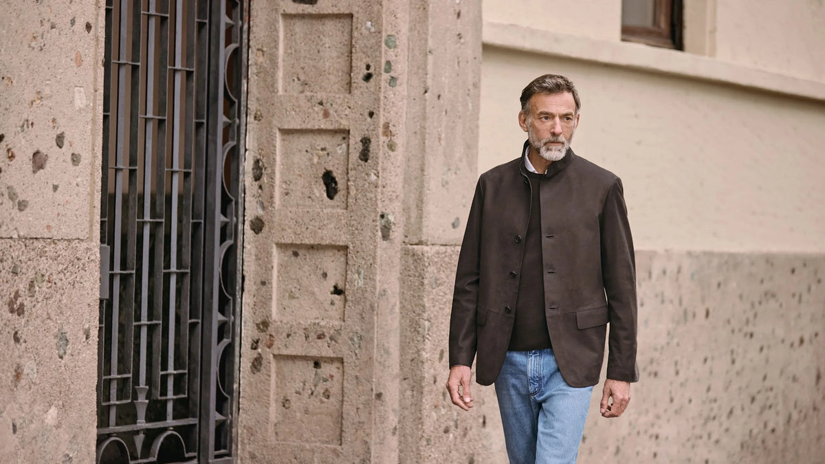 Man in brown jacket and jeans walking past a stone building