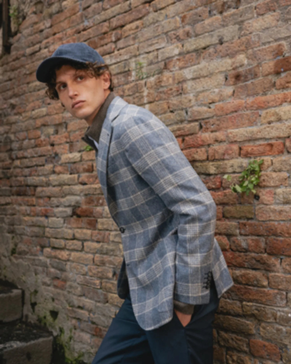 Man in a blue check Barba Napoli jacket and flat cap against a brick wall.
