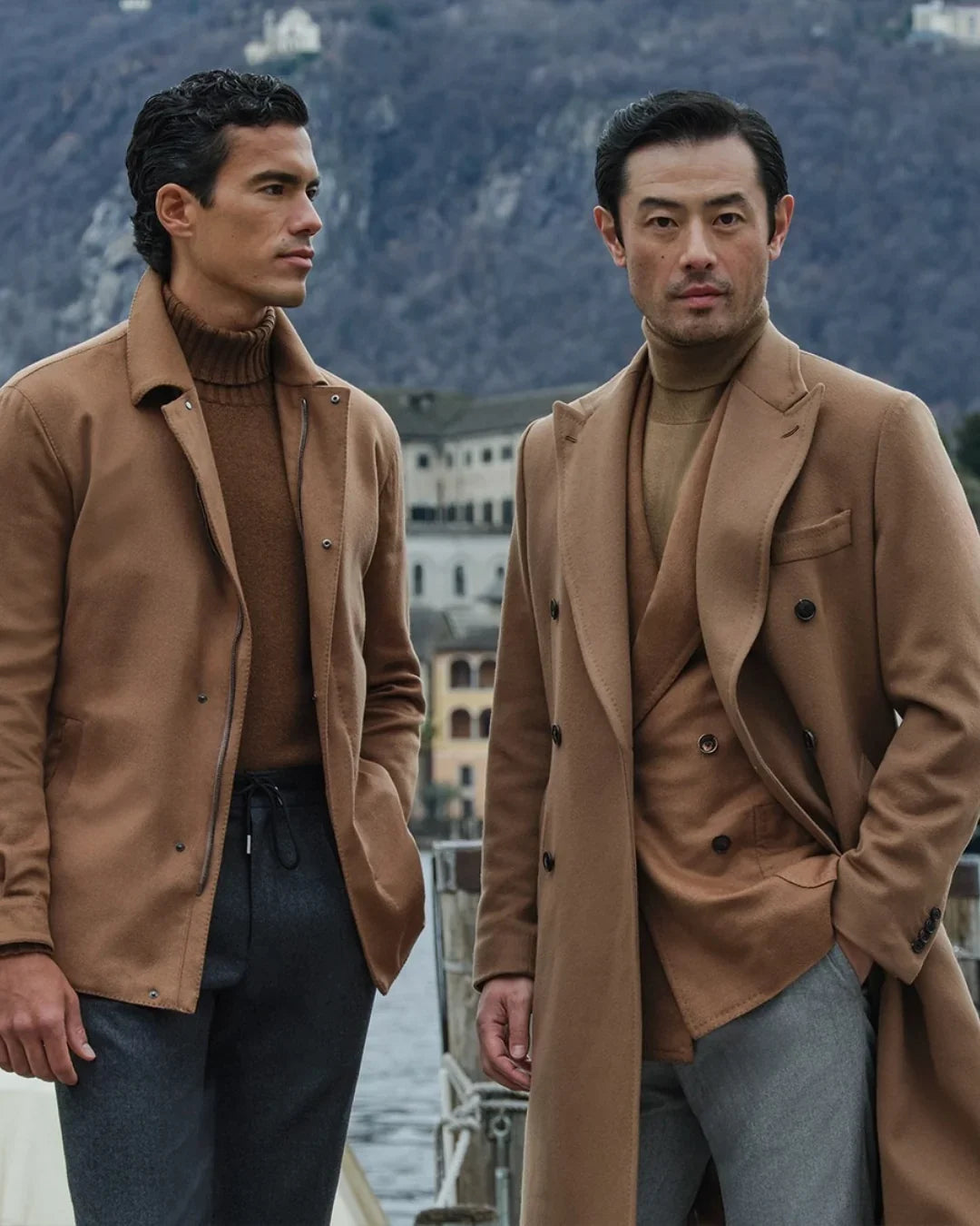 Two men in camel coats and brown turtlenecks by a lake